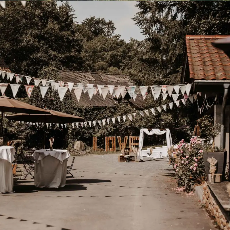 Romantische Hochzeitskulisse im Innenhof der Pension Mausmühle Rustikale Hochzeitsdekoration im Hof der Pension Mausmühle mit LOVE-Schriftzug, Traubogen und Wimpelgirlanden.