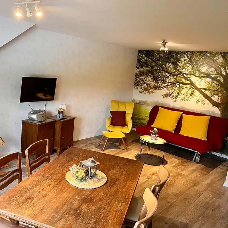 Wohnbereich der Ferienwohnung in der Pension Mausmühle Gemütlich eingerichtetes Wohnzimmer der Ferienwohnung in der Pension Mausmühle mit Naturmotiv-Tapete, Holztisch und farbenfrohen Möbeln.